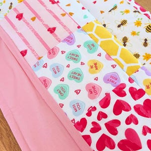 Puede incluir: Una colcha de retazos con varios patrones de tela que incluyen abejas y flores amarillas, rayas rosas con corazones rojos, un estampado de panal amarillo y corazones de caramelo con textos como Be Mine y Miss You impresos en ellos.