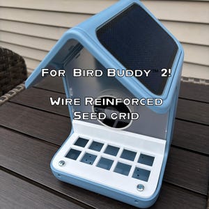 May include: A light blue bird feeder with a dark gray top and a white seed grid. The text "For Bird Buddy 2!" and "Wire Reinforced Seed Grid" are visible on the feeder. The feeder is designed to attract birds.