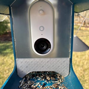 May include: A white security camera mounted on a blue bird feeder with a clear plastic cover. The feeder is filled with birdseed.
