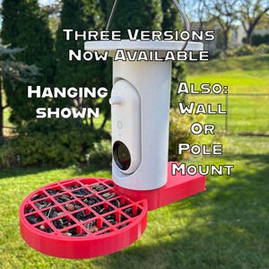 May include: A white speckled bird feeder with a red circular tray containing seeds and a central camera mount. Text reads: THREE VERSIONS NOW AVAILABLE, HANGING SHOWN, ALSO: WALL OR POLE MOUNT. The device features a black lens and a white cylindrical body.