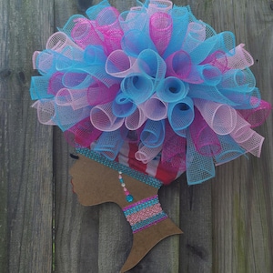 May include: A decorative wreath featuring a silhouette of a woman's head with a vibrant, multi-colored mesh afro. The afro is composed of blue, pink, and light pink mesh. The head is adorned with a patterned head wrap and decorative jewelry.