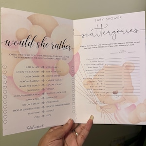 Op de afbeelding: Een spelboekje voor een babyshower met twee pagina's. De linkerpagina heeft een "Would She Rather"-spel met verschillende keuzes. De rechterpagina heeft een "Scattergories"-spel met babygerelateerde categorieën. Het boekje is versierd met roze en perzik kleurige ballonnen.