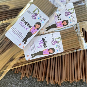 May include: Three packages of incense sticks with labels that say "Dragons Blood", "Indigo Sky", and "Amber Moon". The labels feature a cartoon woman with a purple shirt and black pants. The incense sticks are brown and are packaged in clear plastic.