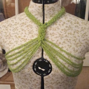 May include: A light green, hand-knitted halter top displayed on a dress form. The halter top features a braided neck strap and multiple cascading strands across the chest. The dress form is covered in a floral fabric.