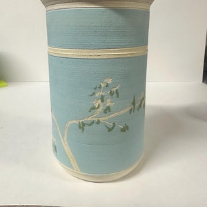 May include: A light blue ceramic vase with a white rim and two white bands. The vase features a green and white floral design.