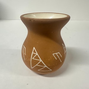 Può includere: Un piccolo vaso di terracotta con una glassa bianca e disegni geometrici. Il vaso ha un corpo arrotondato e un'apertura leggermente svasata.