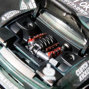 Peut inclure: Une voiture miniature vert foncé avec le capot ouvert révélant un compartiment moteur détaillé doté d'un collecteur d'admission noir et de couvre-culasses rouges. La carrosserie est ornée de décalcomanies blanches dont le texte DRIFT OR DIE.
