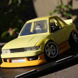 Könnte beinhalten: Ein gelb-oranges Modellauto mit tiefergelegtem Fahrwerk, breiten silbernen Rädern, einer Frontstoßstange mit sichtbarem silbernen Ladeluftkühler und schwarzen Seitenspiegeln. Das Fahrzeug hat eine zweifarbige Lackierung in hellem Gelb und Orange.