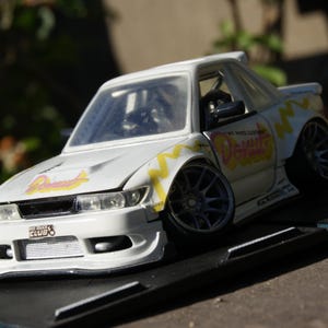 Può includere: Un modellino di auto da drift bianco con kit carrozzeria largo, pneumatici a profilo ribassato e cerchi grigio scuro a razze multiple. Il veicolo presenta grafiche gialle a zigzag, la scritta Donut sul cofano e sulla fiancata e un logo Donut Club.