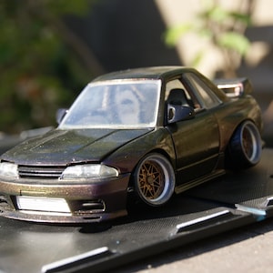 Peut inclure: Un modèle réduit de coupé Nissan Skyline GT-R R32 de couleur marron métallisé foncé. La voiture présente une suspension abaissée, des ailes larges, des jantes dorées à rayons multiples avec des bords argentés, une prise d'air avant et un aileron.