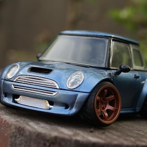 Op de afbeelding: Een blauwe gegoten speelgoedauto met een wide-body kit, een zilveren grille en opvallende koperkleurige wielen. Het voertuig heeft een compact ontwerp met een zwart dak, heldere koplampen en een zichtbare zilveren intercooler.