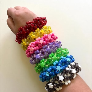 Puede incluir: Una pila de pulseras de cuentas de colores con dijes en forma de estrella. Las pulseras son rojas, amarillas, rosadas, moradas, verdes, azules y negras.