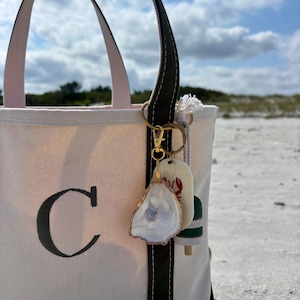 Op de afbeelding: Crème canvas draagtas met een zwarte letter 'C' op de voorkant. De tas heeft donkergroene accenten en een gouden sleutelhanger met oesterschelp- en kreeftenboei-bedels. Achtergrond is een zandstrand onder een blauwe lucht.