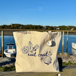 May include: Cream-colored tote bag with blue illustrations of seashells, a lobster, and the words "east coast." A decorative oyster shell charm hangs from the handle. The bag is on a wooden surface with a waterfront background.