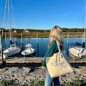 Pode incluir: Uma sacola de lona bege com as palavras "east coast" e um design floral e de conchas marinhas em azul. A sacola &eacute; carregada por uma pessoa em um cais, com veleiros ao fundo.