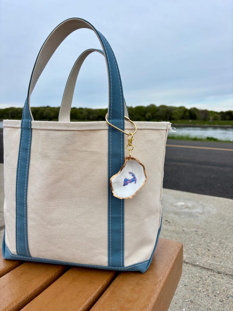 May include: A beige canvas tote bag with blue accents and handles. A gold-colored keychain with an oyster shell charm is attached to the bag. The charm features a painted image of a coastal outline in blue and purple.