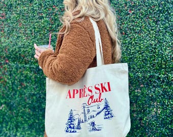 以下が含まれることがあります： 赤と青で「APRÈS SKI Club」の文字が入ったクリーム色のトートバッグ。スキーヤー、木、リフトのある山の風景がデザインされています。バッグには長いオフホワイトのストラップが付いています。