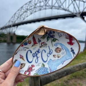 以下が含まれることがあります： 「ケープコッド」の文字、地図、ロブスター、灯台、アイスクリームコーンのイラストが描かれた手描きの装飾的な貝殻の皿。 皿は橋の前で持たれています。