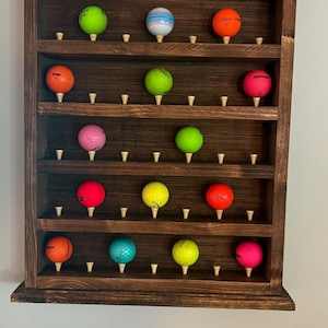 May include: A dark brown wooden wall-mounted display rack with five horizontal shelves. Each shelf features small wooden pegs holding various colored golf balls, including neon green, orange, pink, yellow, and blue, arranged in a grid pattern.
