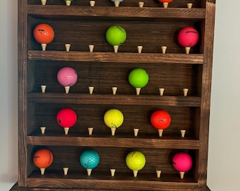 May include: A dark brown wooden wall-mounted display rack with five horizontal shelves. Each shelf features small wooden pegs holding various colored golf balls, including neon green, orange, pink, yellow, and blue, arranged in a grid pattern.