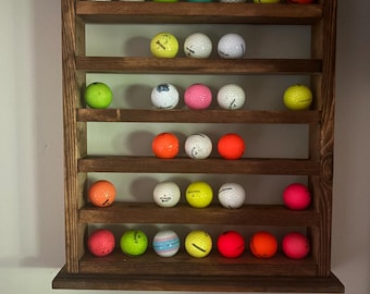 May include: A rectangular wooden display rack with six horizontal shelves holding a collection of multi-colored golf balls. The rack features a dark brown stained finish and is mounted on a wall, showcasing various white, yellow, orange, pink, green, and blue golf balls arranged across the shelves.