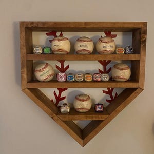 Puede incluir: Una estantería de madera con forma de bate de béisbol con dos estantes. Los estantes están llenos de pelotas de béisbol y anillos con temática de béisbol. El fondo es una pared blanca con un patrón rojo y blanco.