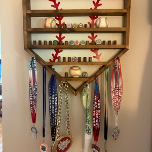 May include: A home plate-shaped wooden wall display rack with horizontal shelves and pegs for hanging medals. The back panel features a red baseball stitch graphic. The shelves hold baseballs and small rings, while various medals hang below.