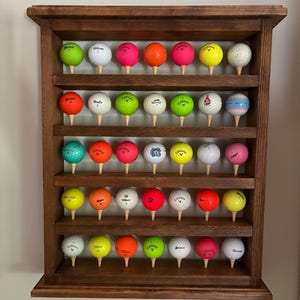 Pode incluir: Um expositor de madeira escura cheio de bolas de golfe de várias cores, incluindo verde, rosa, laranja e branco. Cada bola repousa sobre um tee de madeira. O expositor está montado numa parede de cor clara.