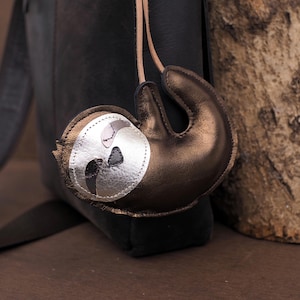 May include: A brown leather sloth-shaped keychain with a silver face and tan leather strap. The sloth is hanging on a black leather backpack. The keychain has a metallic sheen and detailed stitching.