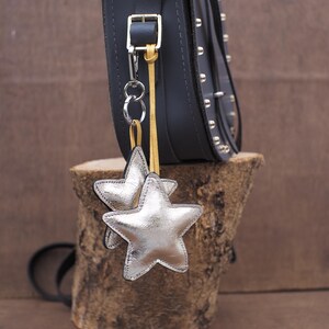 May include: A black leather bag with silver star-shaped charms hanging from a silver chain. The bag has silver studs and a buckle closure. The bag is sitting on a piece of wood.