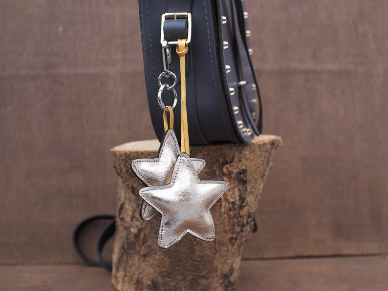 May include: A black leather bag with silver star-shaped charms hanging from a silver chain. The bag has silver studs and a buckle closure. The bag is sitting on a piece of wood.