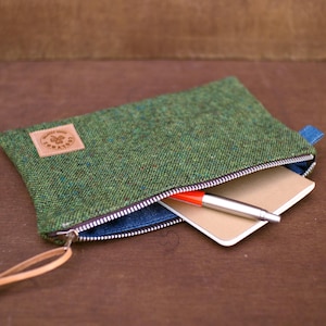 Puede incluir: Bolsa con cremallera de tweed verde con un tirador de cuero y un pequeño parche de cuero. La bolsa está parcialmente abierta, revelando un pequeño cuaderno y un bolígrafo con punta roja. El interior es azul.