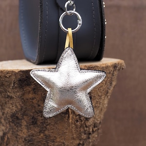 May include: A silver star-shaped keychain with a black border hangs from a silver ring attached to a black purse. The star is made of shiny, textured material. A gold-colored strap connects the star to the ring. The purse is dark gray.