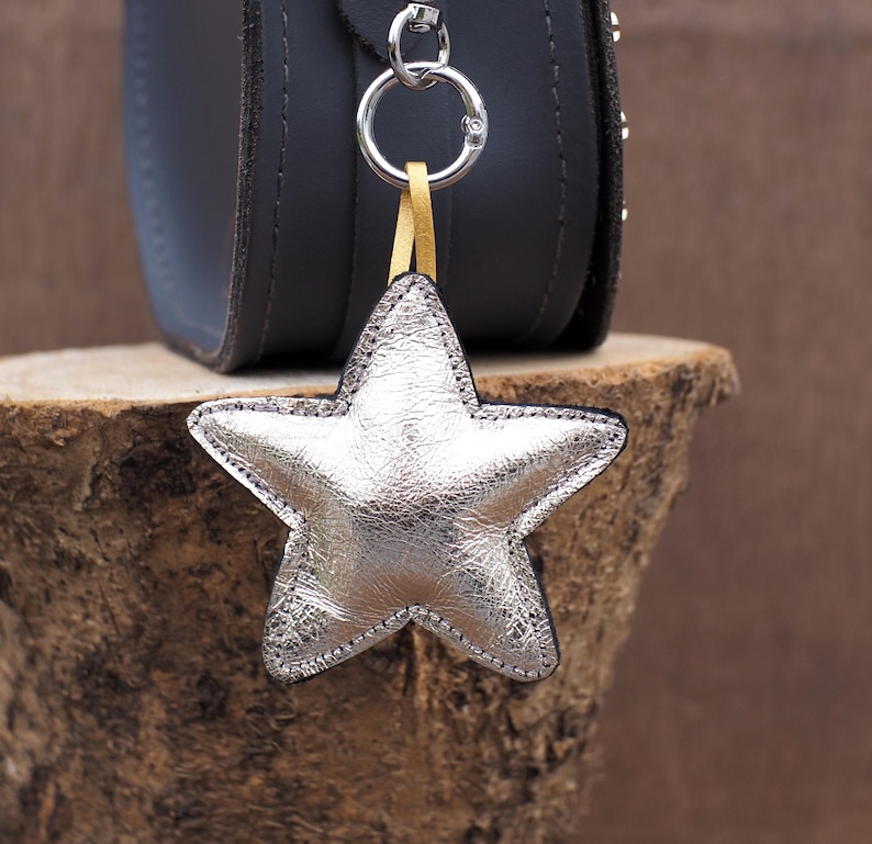 May include: A silver star-shaped keychain with a black border hangs from a silver ring attached to a black purse. The star is made of shiny, textured material. A gold-colored strap connects the star to the ring. The purse is dark gray.