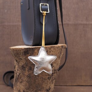 May include: A dark gray leather satchel bag with a silver star-shaped charm hanging from a gold strap. The bag is on a wooden stump, with a dark strap. The star charm is silver and has a stitched outline.