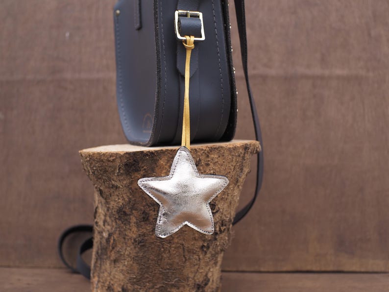 May include: A dark gray leather satchel bag with a silver star-shaped charm hanging from a gold strap. The bag is on a wooden stump, with a dark strap. The star charm is silver and has a stitched outline.