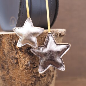 May include: Two silver star-shaped ornaments with gold ribbons hang in front of a dark gray object. The stars are made of a shiny, metallic material and are stitched around the edges. The background is a wooden surface.