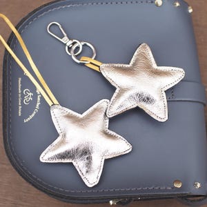May include: Two silver star-shaped keychains with yellow straps and silver hardware. The keychains are resting on a gray leather satchel bag. The bag has a buckle closure and a shoulder strap. The bag is embossed with the text "Handmade in Great Britain".
