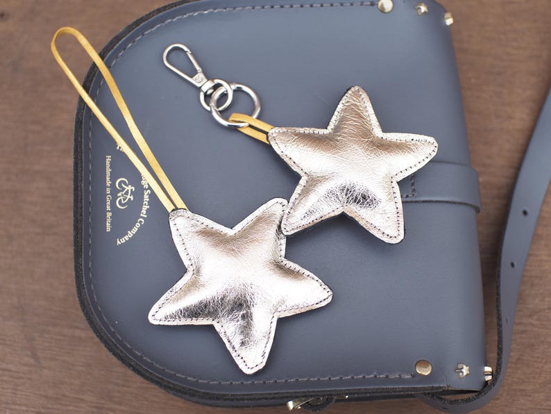 May include: Two silver star-shaped keychains with yellow straps and silver hardware. The keychains are resting on a gray leather satchel bag. The bag has a buckle closure and a shoulder strap. The bag is embossed with the text "Handmade in Great Britain".