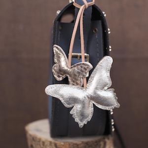 May include: A dark blue handbag with silver butterfly embellishments. The bag has a top handle and a strap. The butterflies are made of a metallic silver material and are attached to the bag with a leather cord.