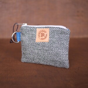 Könnte beinhalten: Eine kleine graue und schwarze Reißverschlusstasche mit blau-weißem Futter. Die Tasche hat einen silbernen Ring und ein braunes Leder-Patch mit dem Text "COOLTHOE HAND CRAFTED" und einem vierblättrigen Kleeblatt.