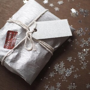 May include: A wrapped gift in silver paper tied with twine, featuring a blank tag and a sticker that reads "COOLYROE HANDCRAFTED IRELAND". Silver star-shaped confetti is scattered on the wooden surface.