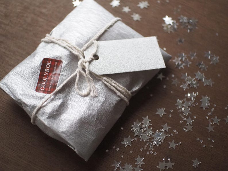 May include: A wrapped gift in silver paper tied with twine, featuring a blank tag and a sticker that reads "COOLYROE HANDCRAFTED IRELAND". Silver star-shaped confetti is scattered on the wooden surface.
