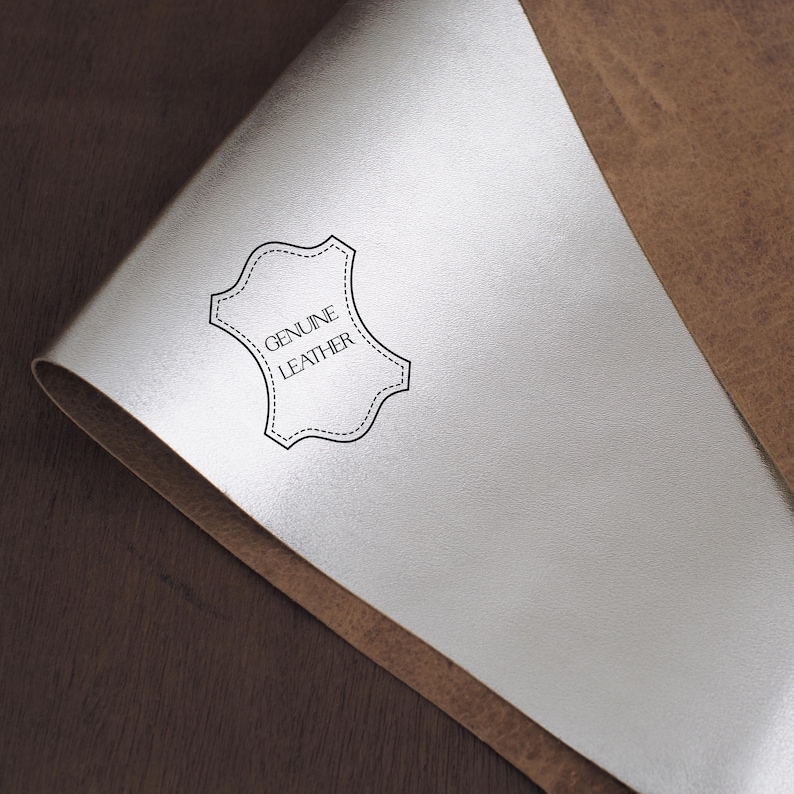 May include: A close-up of a piece of light gray leather with a "GENUINE LEATHER" label. The label is a black outline of a shield shape with a dotted border. The leather is folded over, revealing a brown leather surface.