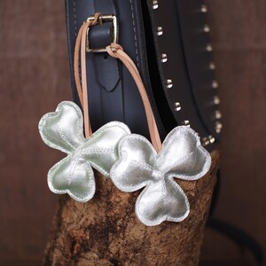 May include: Two silver-colored, clover-shaped charms with leather straps hang near a dark blue handbag. The charms have a metallic sheen and are attached to the bag with tan leather straps. The bag has a buckle and silver studs.