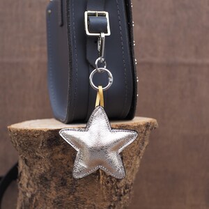 May include: A silver star-shaped leather bag charm hanging from a dark gray crossbody bag. The charm is attached with a silver ring and a yellow strap. The bag is studded with small silver rivets.