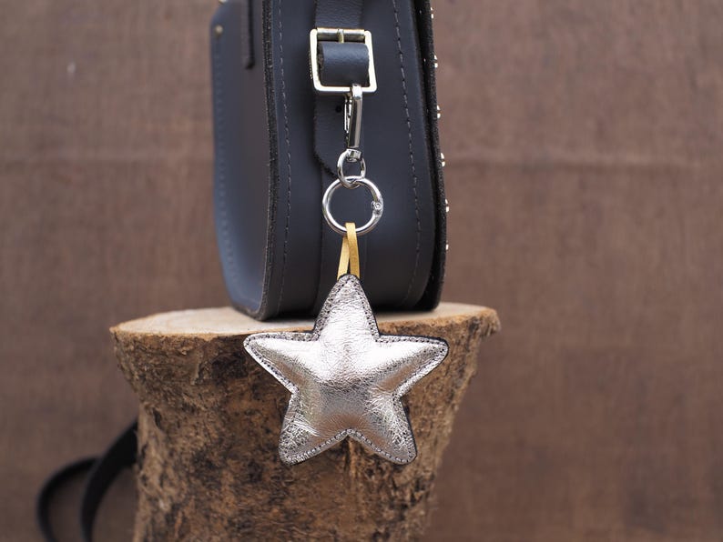 May include: A silver star-shaped leather bag charm hanging from a dark gray crossbody bag. The charm is attached with a silver ring and a yellow strap. The bag is studded with small silver rivets.