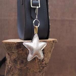 May include: A silver star-shaped charm hangs from a dark gray leather bag. The charm is attached with a silver clasp and a yellow strap. The bag is on a wooden surface.