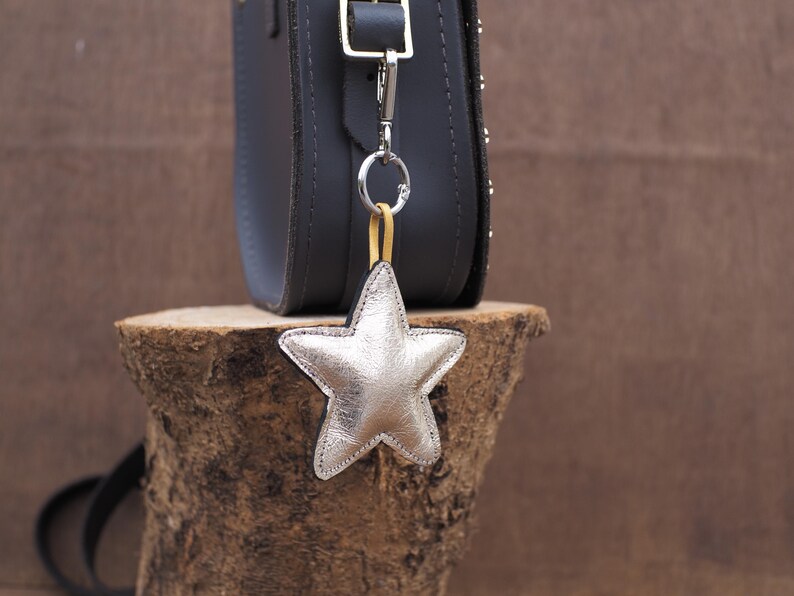 May include: A silver star-shaped charm hangs from a dark gray leather bag. The charm is attached with a silver clasp and a yellow strap. The bag is on a wooden surface.