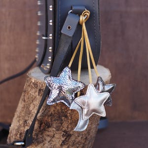 May include: A dark gray leather crossbody bag with silver studs and star-shaped charms. The charms are silver and iridescent, hanging from gold-colored cords. The bag is displayed on a wooden log against a brown background.